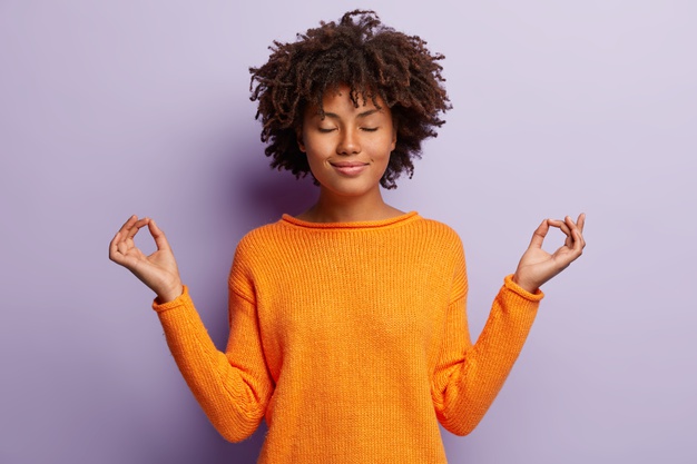 mulher de aparencia agradavel e calma medita dentro de casa segura as maos em gesto de mudra tem um sorriso encantador olhos fechados usa roupas laranja modelos sobre parede roxa