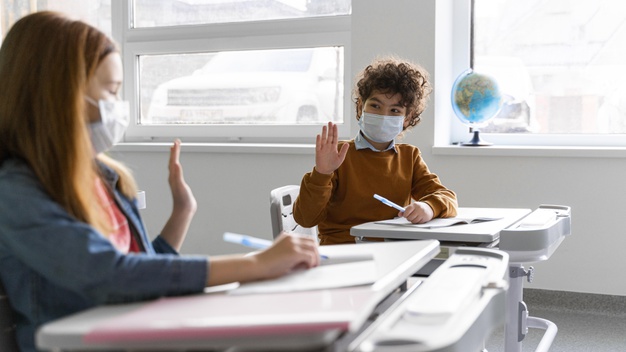 vista lateral de criancas com mascaras medicas em sala de aula saudando se a distancia