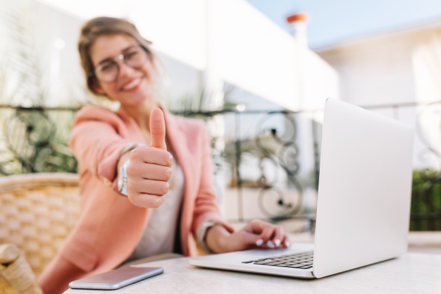 mulher jovem bonita estudante mulher de negocios mostrando os polegares muito bem sentado no cafe ao ar livre no terraco com o laptop vestindo roupas rosa inteligentes