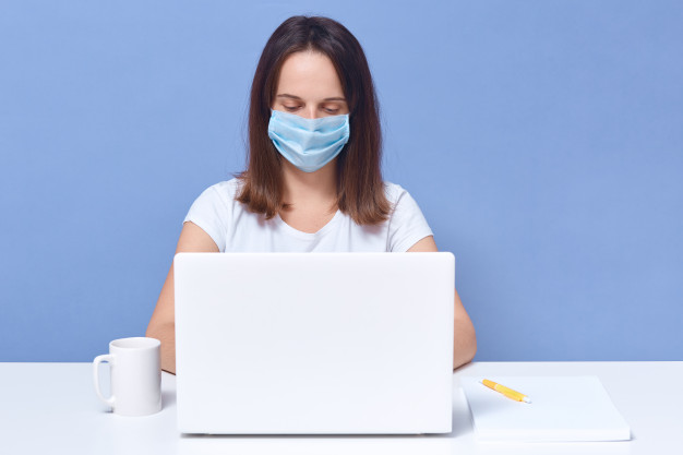 senhora de cabelos escura vestindo camiseta casual e mascara medica que trabalha na frente da tela do colo superior parece concentrado femea estudante fazendo tarefa universitaria on