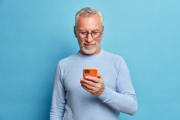 homem barbudo satisfeito focado em smart phone surfa internet envia mensagens de texto em redes sociais usa tecnologias modernas usa jaqueta azul casual poses indoor