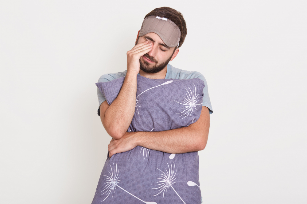 sleepy bearded male posing with closed eyes isolated over white wall rubbing his eyes with hand embracing pillow