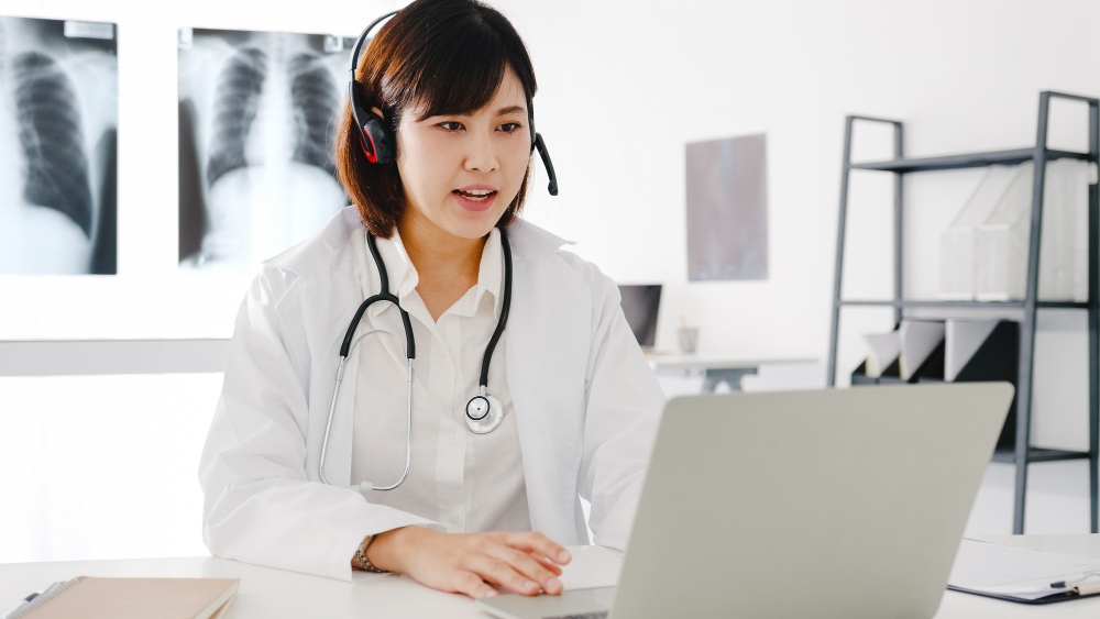 young asia female doctor in white medical uniform with stethoscope using computer laptop talking video conference call with patient at desk in health clinic or hospital