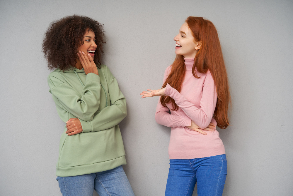 cheerful young lovely ladies smiling positively while meeting each other and having pleasant talk wearing comfortable casual clothes while posing over grey wall