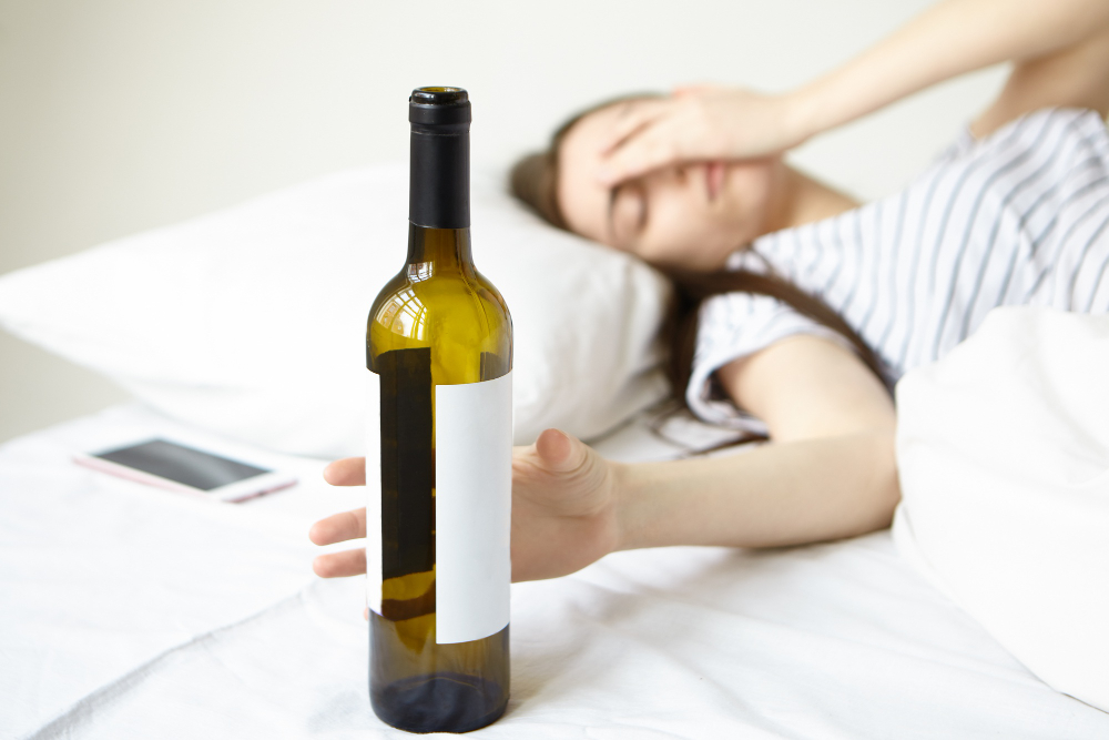 indoor shot of brunette young woman lying in white bed touching face having bad headache after night party waking up in the morning with hangover selective focus on hand holding bottle of wine