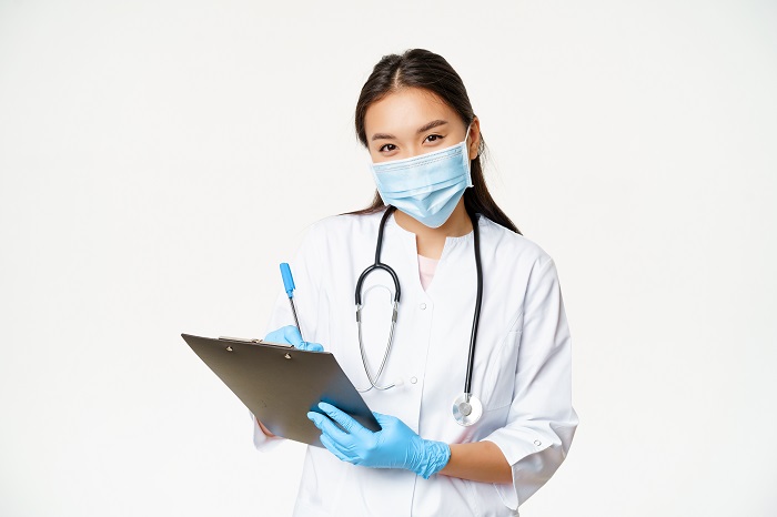 Smiling asian female doctor in face mask and gloves, writing down patient diagnose, prescription, holding pen and clipboard, white background