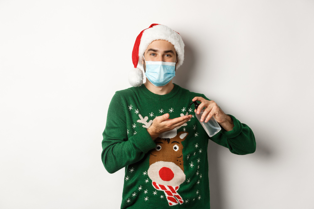 concept of covid and christmas holidays caucasian man in face mask and sweater using antiseptic clean hands with sanitizer standing over white background