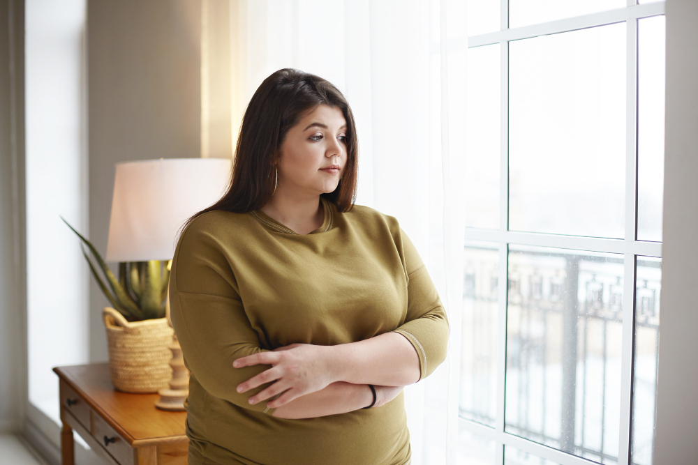people lifestyle and body positivity concept beautiful young brunette female with extra pounds standing by window at home crossing arms on her chest having thoughtful pensive look pondering