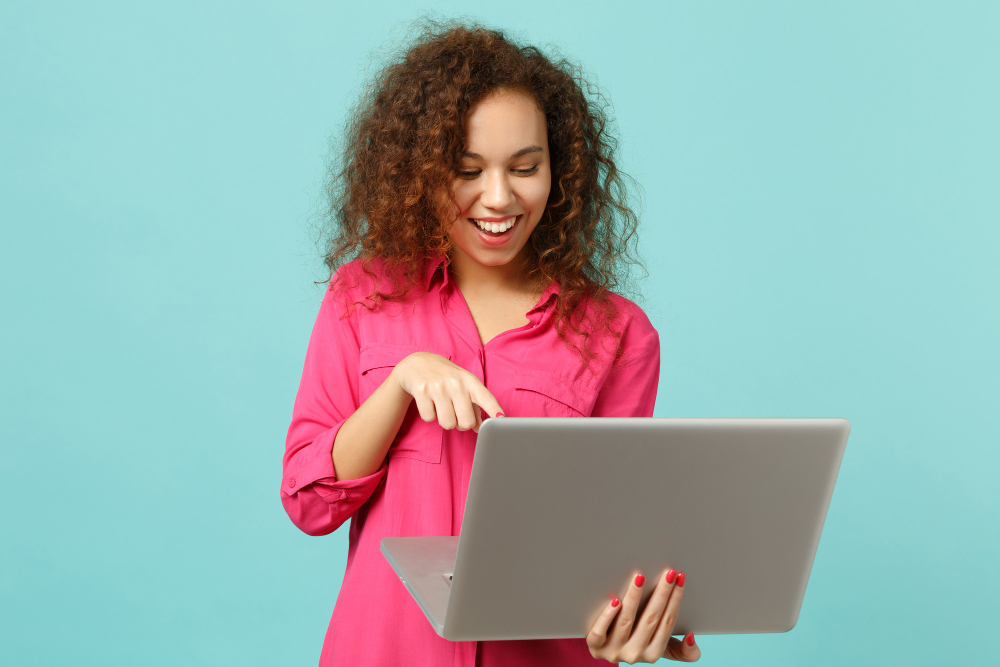 smiling african girl casual clothes pointing index finger laptop pc computer isolated blue turquoise wall background studio people sincere emotions lifestyle concept mock up copy space