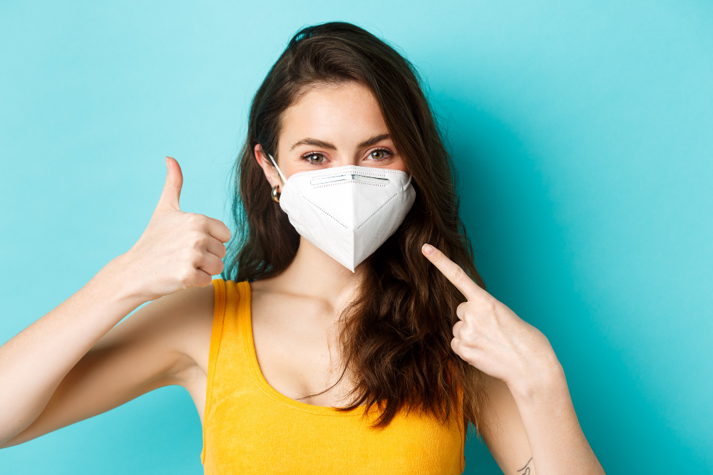 covid coronavirus and social distancing wear face mask smiling woman in medical respirator pointing at face showing thumb up standing over blue background