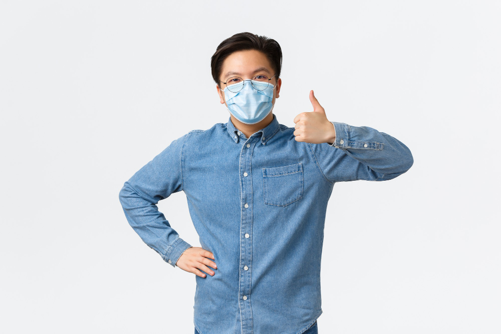 covid preventing virus and social distancing at workplace concept supportive cheerful asian male entrepreneur in medical mask showing thumbs up in approval praise teamwork