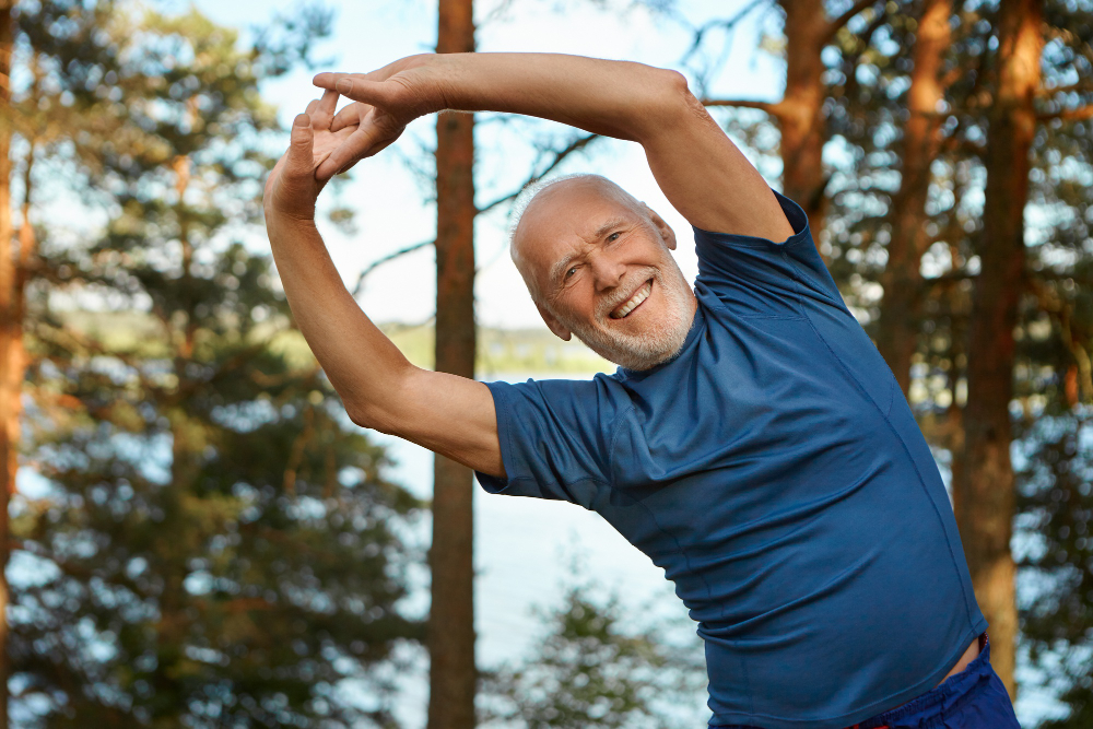outdoor shot of happy energetic senior retired man enjoying physical training in park doing side bends exercise holding hands together with broad smile warming up body before run