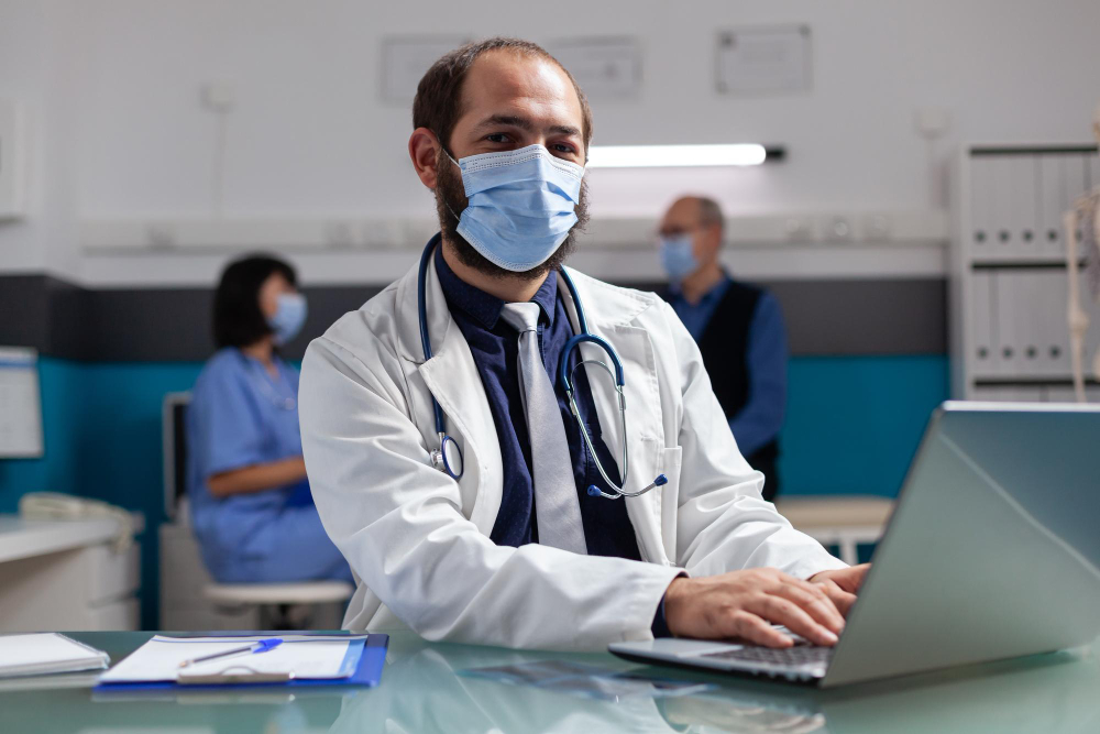 portrait of doctor looking at camera and using computer in office physician with face mask wearing white coat at consultation appointment working with laptop for health care career