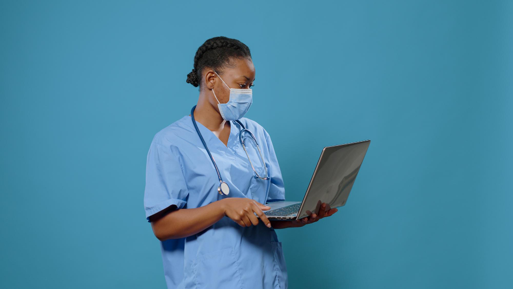 woman nurse wearing face mask and using laptop in front of camera medical assistant in uniform having protection against coronavirus while holding computer to work on healthcare