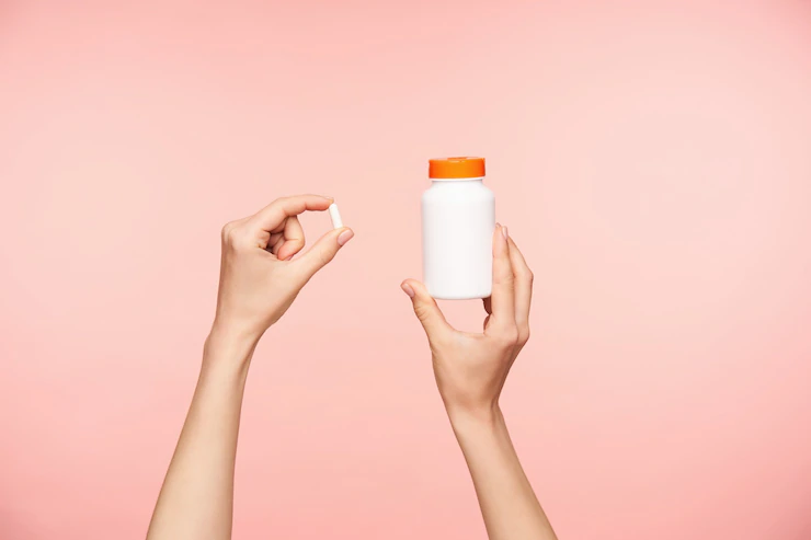 foto recortada das maos bem cuidadas de uma mulher levantada segurando um comprimido branco e um frasco com tampa laranja tomando vitaminas enquanto posava sobre um fundo rosa