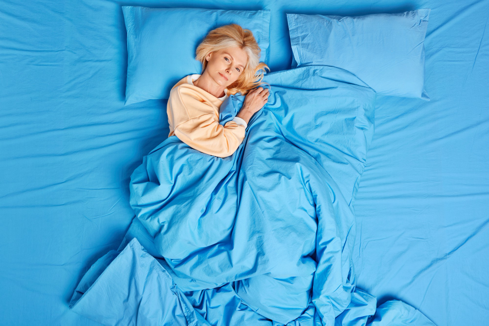calm european middle aged woman awakes satisfied after seeing good dreams poses well slept under blue blanket wears pajama feels comfortable enjoys lazy day bed time and cozy morning concept