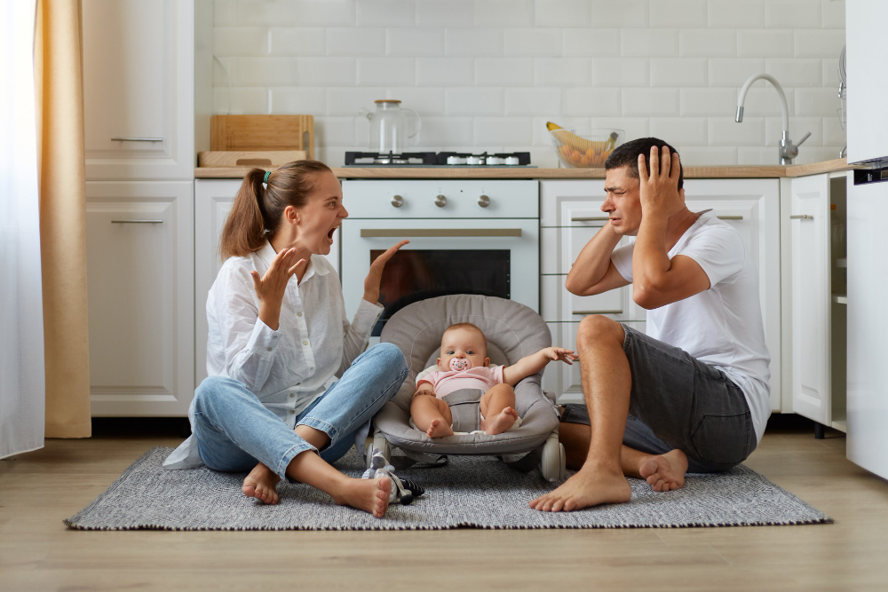 indoor short of arguing couple sitting on the floor in kitchen wife screaming loudly husband covering ears with palms family posing with infant baby in rocking chair