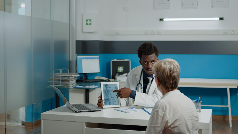 young physiotherapist showing tablet of bones injury to senior patient with osteoporosis at desk in doctors office elder woman looking at virtual vertebrae for medical examination
