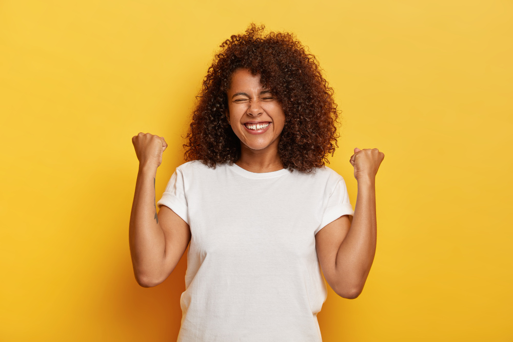 isolated shot of beautiful successful woman with curly hair raises clenched fists celebrates triumph being very pleased and happy closes eyes from pleasure wears white t shirt yeah i did it