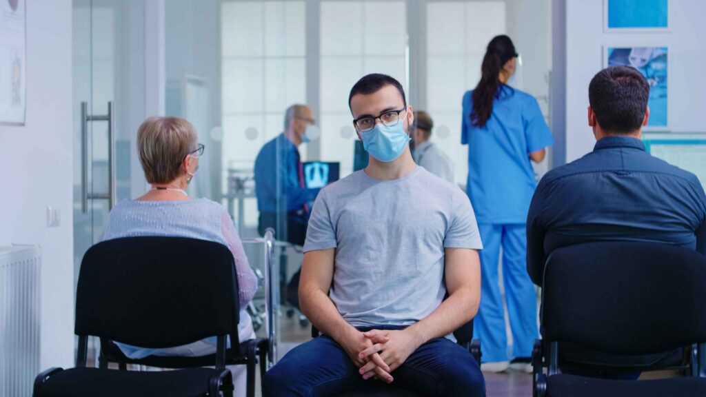 young man with face mask against coronavirus looking at camera in hospital waiting area senior woman with walking frame waiting for consultation in clinic
