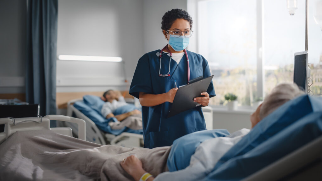 Hospital Ward: Friendly Nurse Wearing Face Mask Talks to Beautiful Senior Female Patient Resting in Bed, Checks Her Vitals, Writes down Information Head Nurse and Old Lady Recovering after Covid